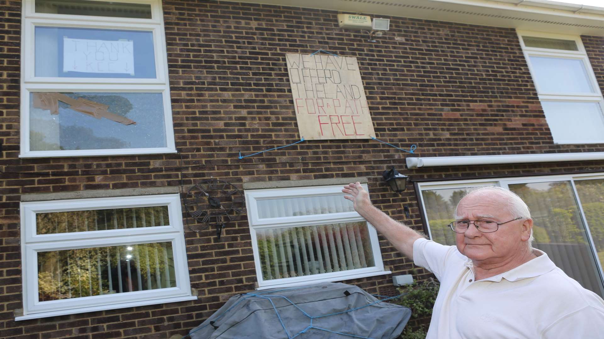 Keith Endacott points to a broken window and signs letting the public know that he is not against a pathway along the rear of his house