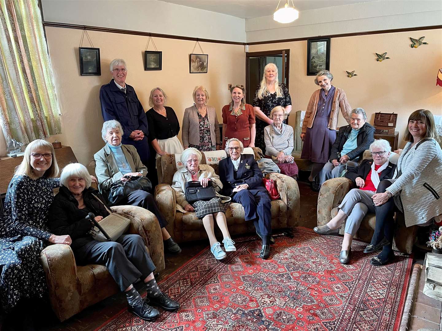 Iris and May seated together on the sofa surrounded by Inspired Friendships members and volunteers of the Old Forge Wartime House