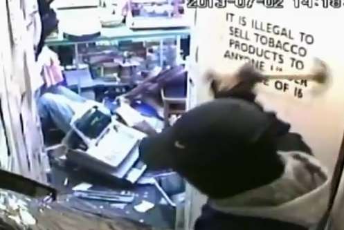 A robber uses a hammer to smash the glass again - while the staff members lies on the ground with a till on top of her