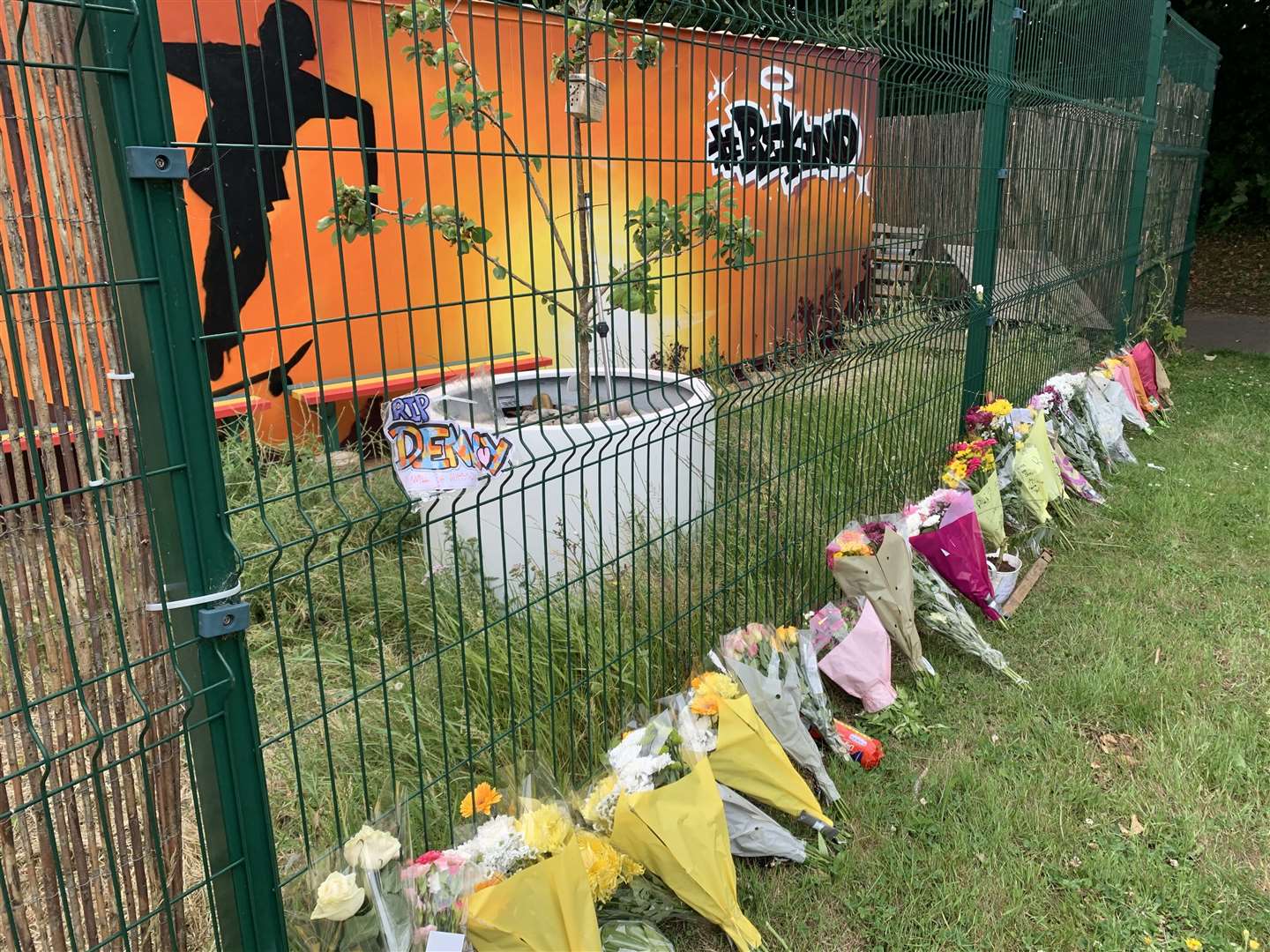 Floral tributes line a grass bank at Deal Skatepark in front of colourful artwork which reads 'Be Kind'