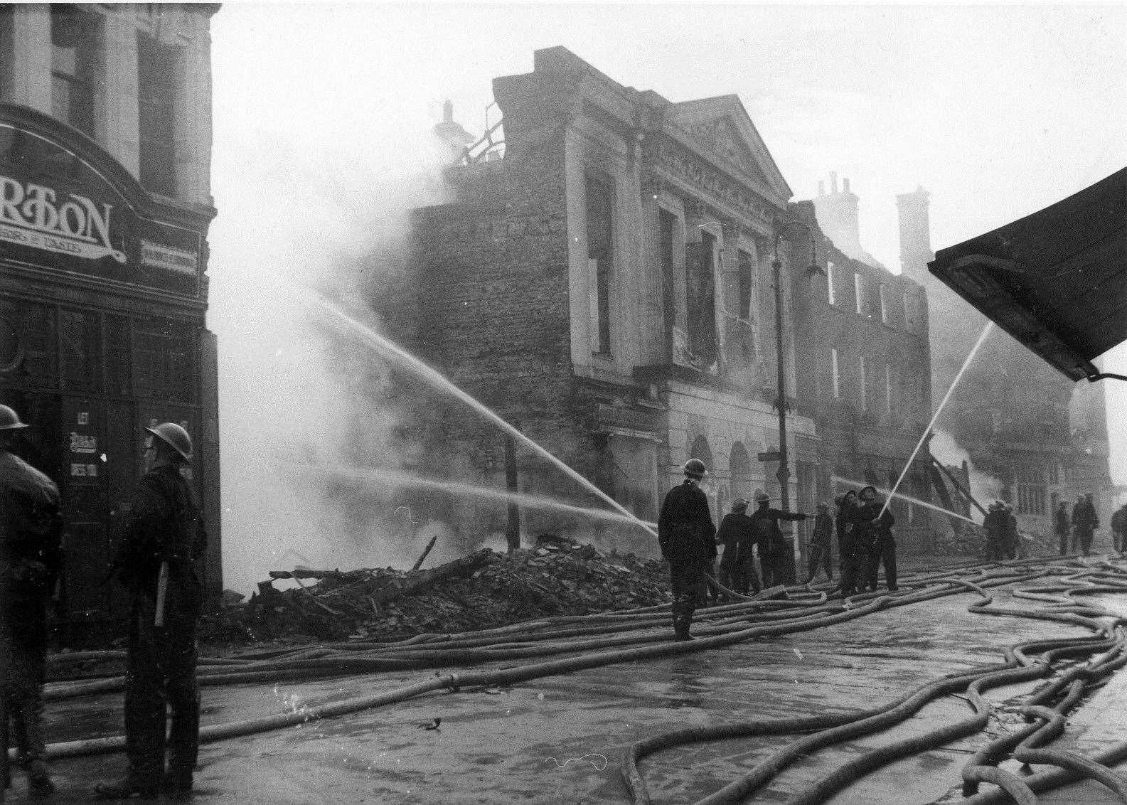 Firemen damp down the remains of buildings on boths sides of St George's Street after the June 1942 Blitz of Canterbury. The imposing Regency facade of the Corn Exchange/Longmarket building remained intact