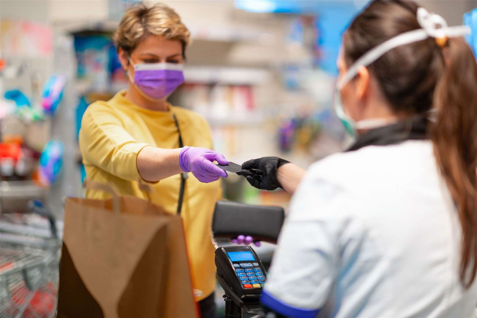 Cashier returning credit card at the cash register to woman with wallet wearing protective face mask and gloves to prevent viruses
